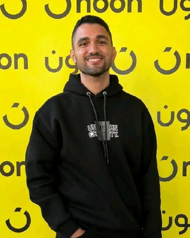 Mohamed smiling in a black hoodie in front of a yellow Noon logo wall